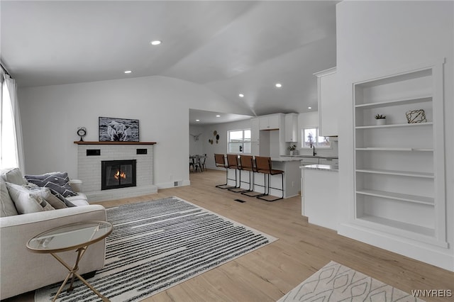 living room with vaulted ceiling, built in features, sink, a brick fireplace, and light wood-type flooring