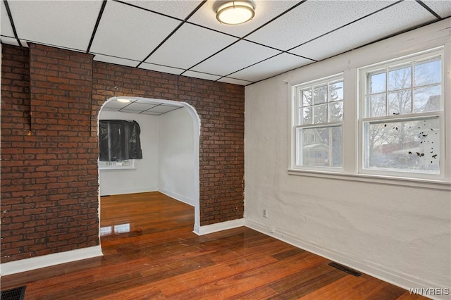 unfurnished room with brick wall, dark wood-type flooring, and a drop ceiling