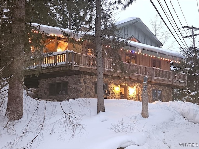 snow covered back of property featuring board and batten siding, stone siding, a balcony, and a wooden deck