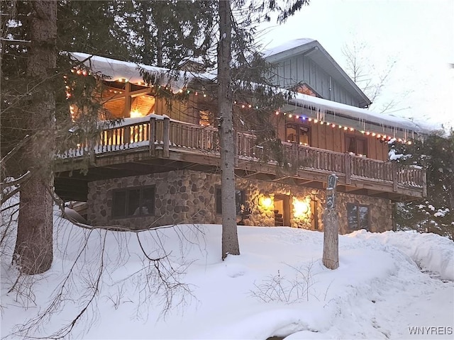 snow covered back of property featuring board and batten siding, stone siding, a balcony, and a wooden deck