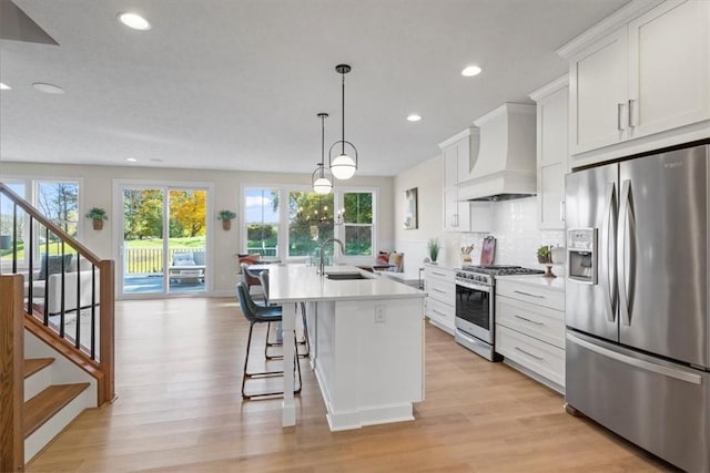 kitchen with appliances with stainless steel finishes, sink, custom range hood, decorative light fixtures, and a kitchen island with sink