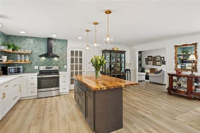 kitchen featuring wooden counters, stainless steel appliances, wall chimney range hood, decorative light fixtures, and white cabinetry