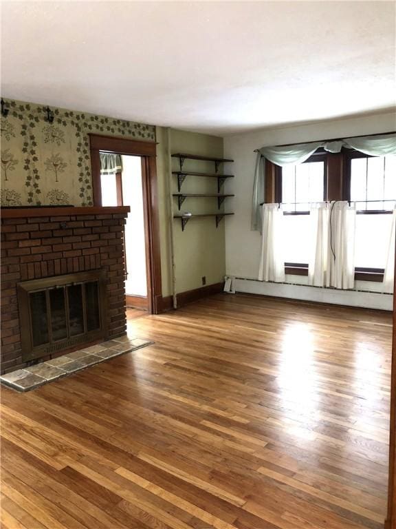 unfurnished living room featuring wallpapered walls, a brick fireplace, wood finished floors, and a wealth of natural light