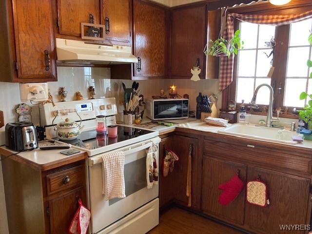 kitchen with white electric range oven, brown cabinets, light countertops, under cabinet range hood, and a sink