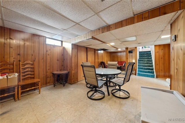 dining space with a drop ceiling, wooden walls, light floors, and stairs