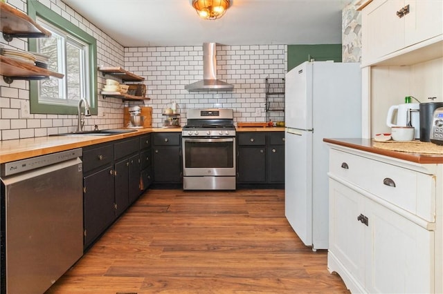 kitchen with open shelves, stainless steel appliances, a sink, dark cabinets, and wall chimney exhaust hood