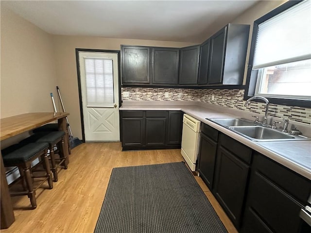 kitchen with light wood finished floors, dark cabinets, white dishwasher, light countertops, and a sink