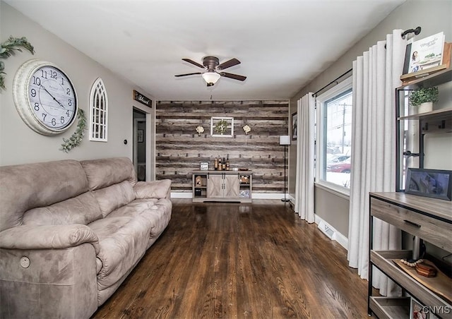 living area with dark wood-type flooring, an accent wall, ceiling fan, wooden walls, and baseboards