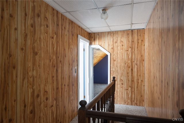 hallway with a paneled ceiling, wooden walls, and an upstairs landing