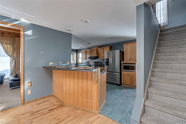 kitchen with a peninsula, light wood-style floors, appliances with stainless steel finishes, wall chimney range hood, and decorative backsplash