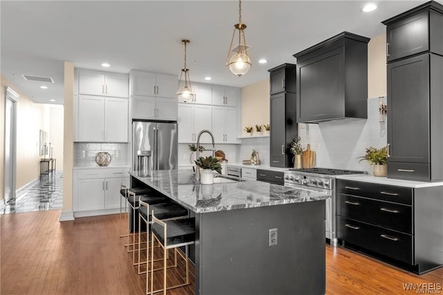 kitchen featuring a center island with sink, high end appliances, wall chimney exhaust hood, decorative light fixtures, and a sink