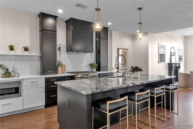 kitchen with dark wood-style flooring, decorative light fixtures, a center island with sink, stainless steel microwave, and a sink