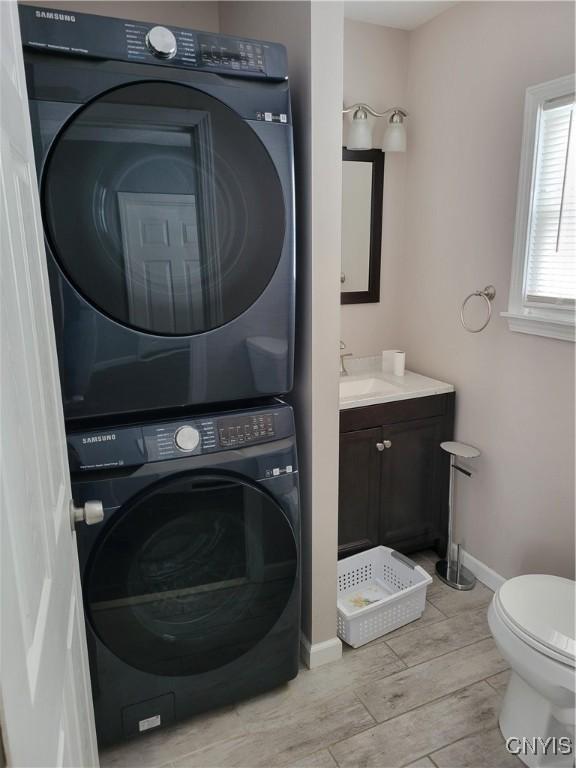 clothes washing area featuring wood finish floors, a sink, stacked washer and clothes dryer, and baseboards