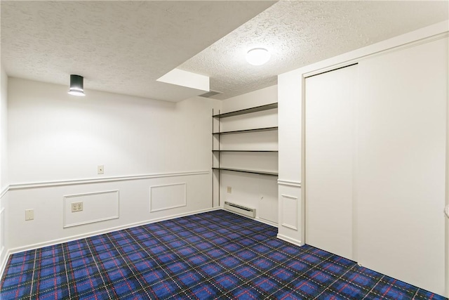 interior space featuring dark colored carpet, a decorative wall, a baseboard heating unit, wainscoting, and a textured ceiling