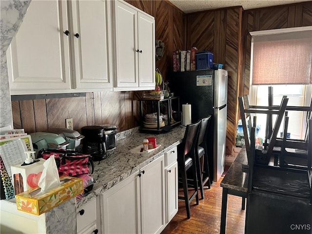 kitchen with white cabinets, dark wood-style floors, freestanding refrigerator, light stone countertops, and wood walls