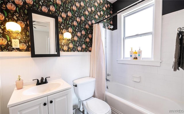 bathroom with a wainscoted wall, toilet, shower / bath combo with shower curtain, vanity, and wallpapered walls