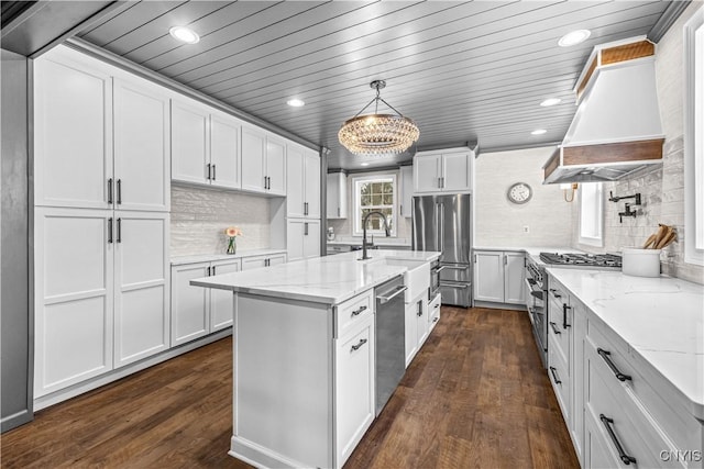 kitchen featuring premium appliances, decorative light fixtures, white cabinets, a center island with sink, and custom range hood