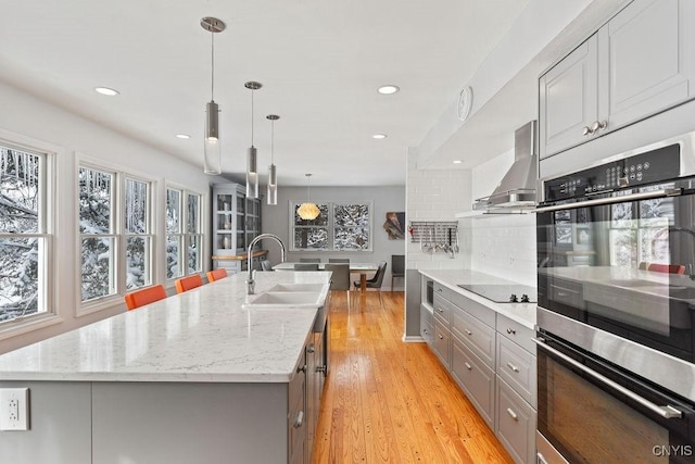 kitchen with pendant lighting, double oven, a kitchen island with sink, a sink, and exhaust hood