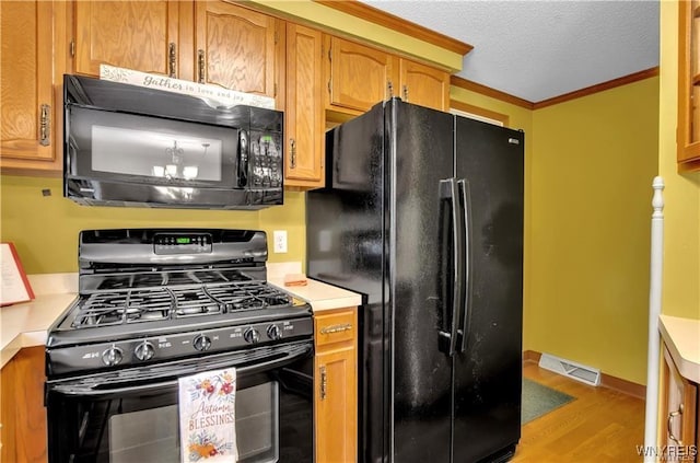 kitchen with light countertops, black appliances, light wood finished floors, brown cabinetry, and crown molding