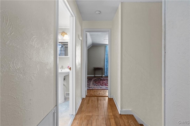 hall with light wood-style floors, vaulted ceiling, a textured wall, and baseboards