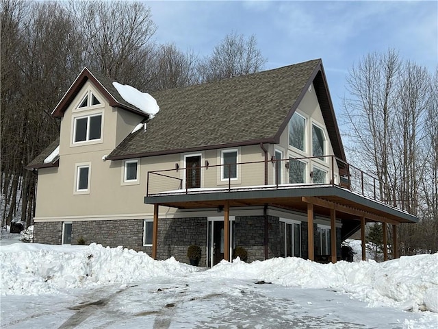 exterior space with a shingled roof, stone siding, a balcony, and stucco siding