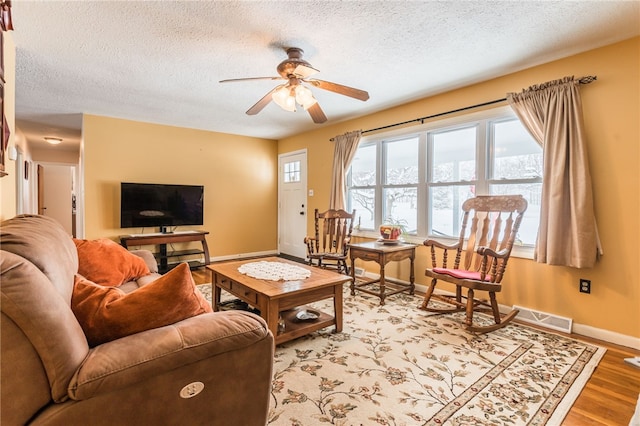 living room featuring a textured ceiling, a ceiling fan, visible vents, baseboards, and light wood finished floors