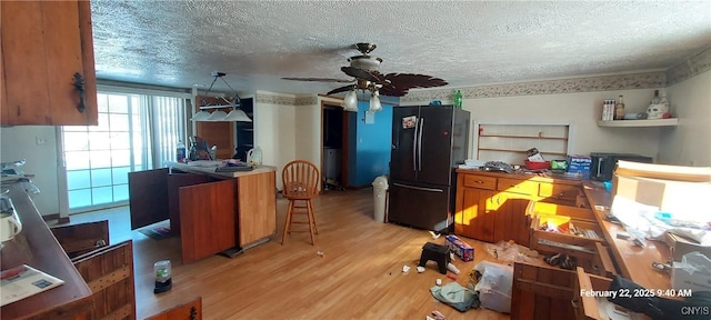 kitchen with a textured ceiling, light wood-style flooring, a ceiling fan, freestanding refrigerator, and brown cabinets