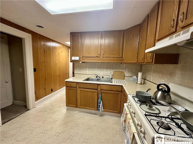 kitchen featuring white range with gas stovetop, brown cabinetry, light countertops, under cabinet range hood, and backsplash