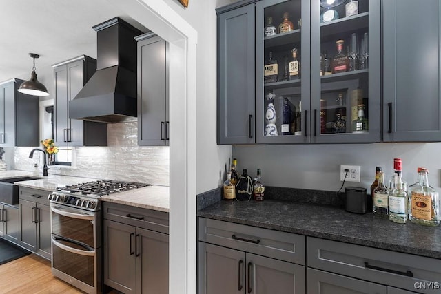 kitchen with range with two ovens, glass insert cabinets, a sink, dark stone countertops, and wall chimney exhaust hood