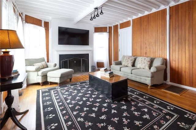 living room featuring wooden walls, a fireplace with flush hearth, beam ceiling, wood-type flooring, and track lighting