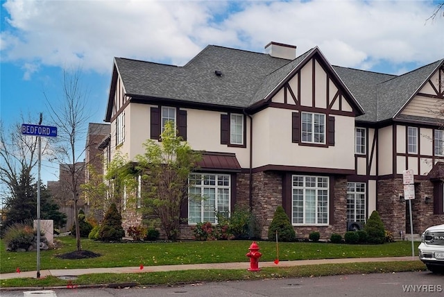 english style home with stucco siding, a front lawn, stone siding, a shingled roof, and a chimney