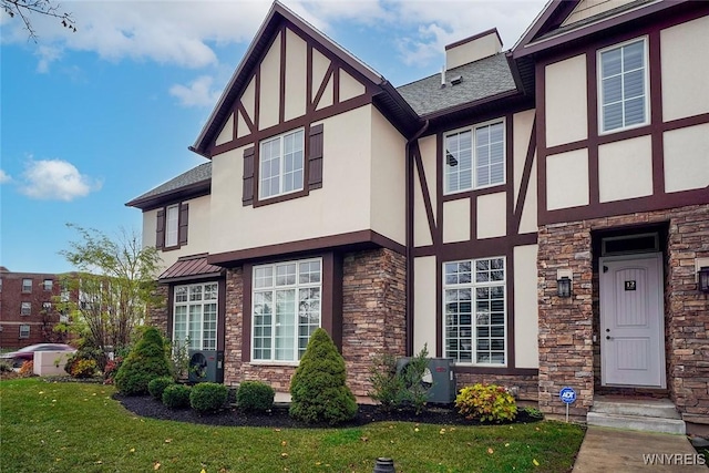 tudor-style house featuring stucco siding, stone siding, roof with shingles, a front yard, and a chimney