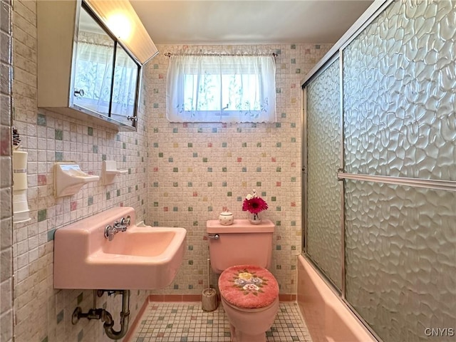 bathroom featuring toilet, bath / shower combo with glass door, tile patterned flooring, and tile walls