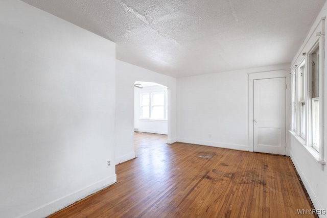 empty room with arched walkways, a textured ceiling, baseboards, and hardwood / wood-style flooring