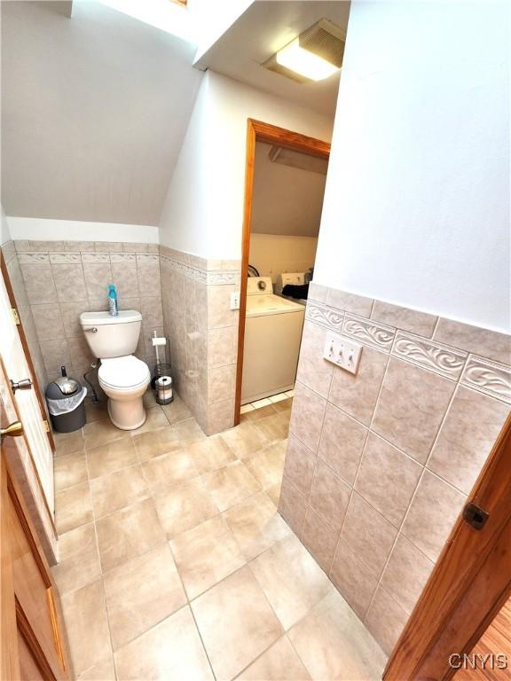 half bath with toilet, a wainscoted wall, separate washer and dryer, tile walls, and vaulted ceiling