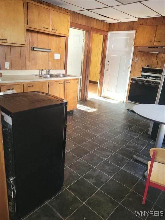 kitchen with a sink, wooden walls, dark tile patterned flooring, white range with gas stovetop, and under cabinet range hood