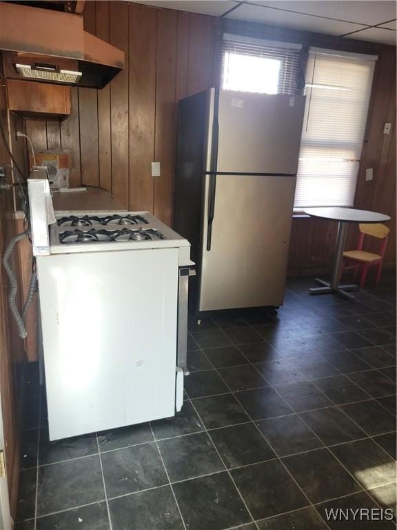 kitchen featuring white gas range oven, brown cabinetry, freestanding refrigerator, wood walls, and under cabinet range hood