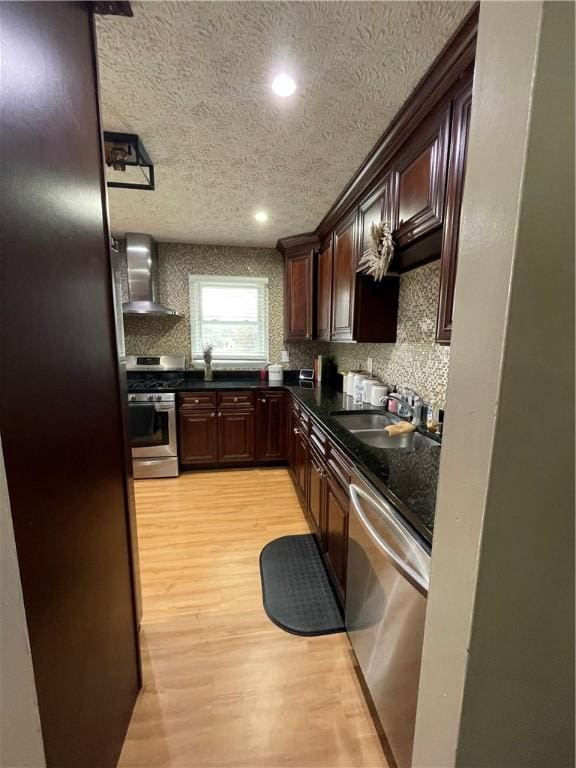 kitchen with light wood-style flooring, stainless steel appliances, a sink, wall chimney exhaust hood, and dark countertops