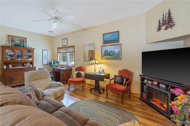 living area featuring ceiling fan, a glass covered fireplace, wood finished floors, and baseboards