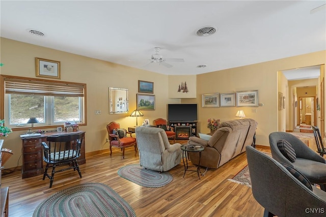 living area featuring a ceiling fan, light wood-type flooring, visible vents, and a glass covered fireplace