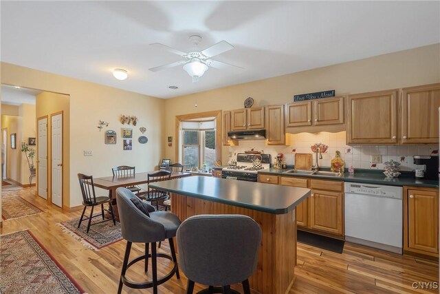 kitchen with under cabinet range hood, white appliances, a sink, light wood-type flooring, and dark countertops