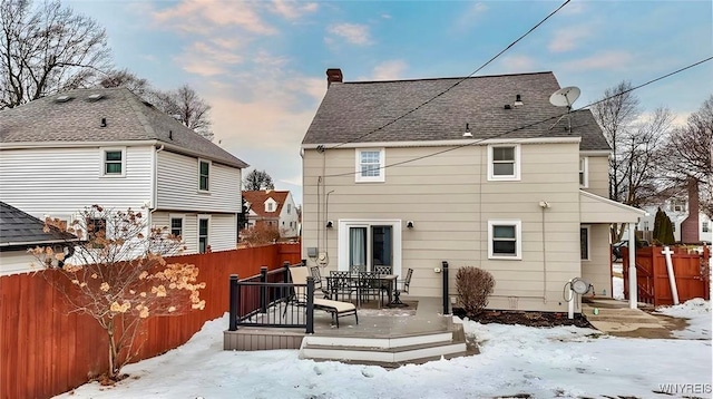 snow covered back of property with a shingled roof, a chimney, a fenced backyard, and a wooden deck