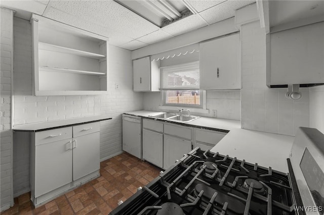 kitchen featuring brick floor, a drop ceiling, white dishwasher, a sink, and open shelves