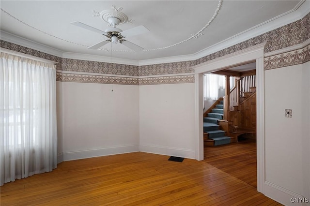 empty room with light wood-style flooring, a ceiling fan, baseboards, ornamental molding, and stairway