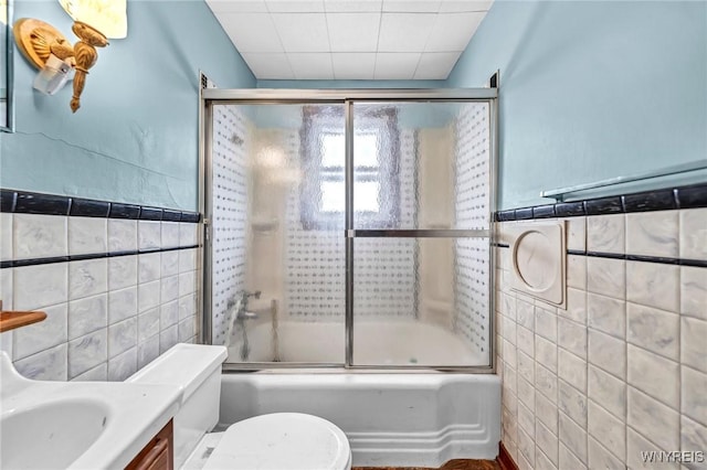 bathroom featuring toilet, bath / shower combo with glass door, vanity, and tile walls