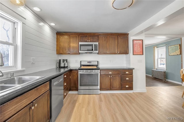 kitchen with stainless steel appliances, light wood-style floors, brown cabinets, radiator heating unit, and dark countertops