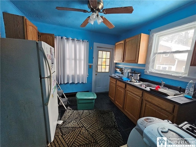 kitchen with ceiling fan, a sink, freestanding refrigerator, brown cabinetry, and dark countertops