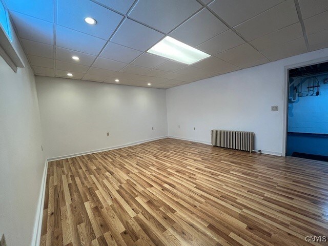 finished basement with baseboards, radiator heating unit, wood finished floors, a paneled ceiling, and recessed lighting