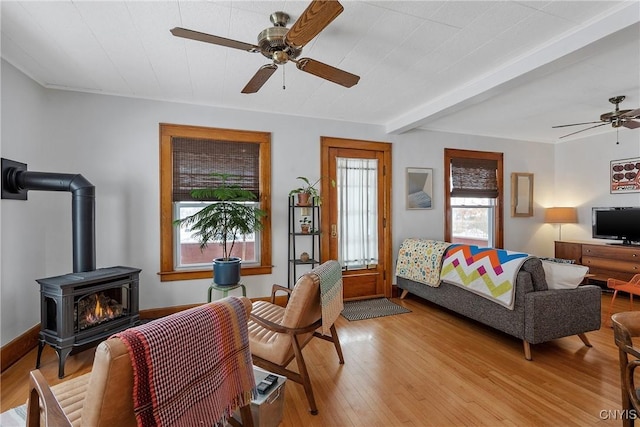 living room featuring light wood finished floors, baseboards, ceiling fan, a wood stove, and beam ceiling