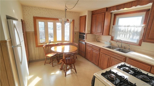 kitchen with a wainscoted wall, brown cabinetry, a sink, white appliances, and wallpapered walls